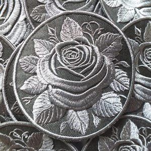 May include: Embroidered patches featuring a detailed rose design in shades of gray. The patches are circular, with the rose and leaves intricately stitched onto a dark gray fabric. The design is repeated across multiple patches.