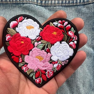 May include: Heart-shaped embroidered patch featuring a floral design. The patch has a black border and is filled with red, white, and pink flowers with green leaves. The patch is held in a hand against a denim background.