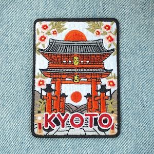 Peut inclure: Écusson brodé représentant un temple japonais avec un soleil orange et le mot "KYOTO" en rouge. Le patch a une bordure noire et des motifs floraux.