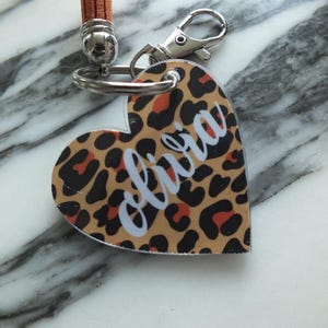 Puede incluir: Llavero en forma de coraz&oacute;n con estampado de leopardo en marr&oacute;n, negro y naranja. El nombre "olivia" est&aacute; escrito en cursiva blanca. Tiene un cierre plateado y una correa de cuero marr&oacute;n.