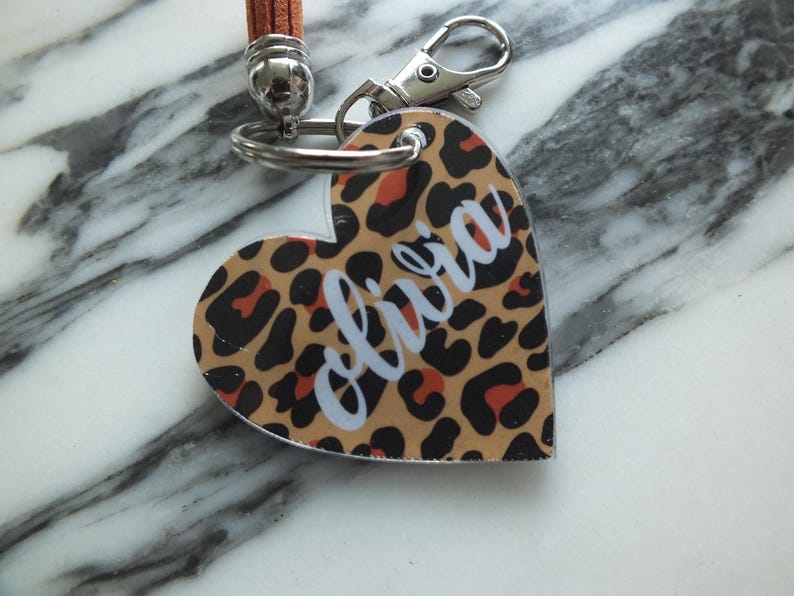 Puede incluir: Llavero en forma de coraz&oacute;n con estampado de leopardo en marr&oacute;n, negro y naranja. El nombre "olivia" est&aacute; escrito en cursiva blanca. Tiene un cierre plateado y una correa de cuero marr&oacute;n.