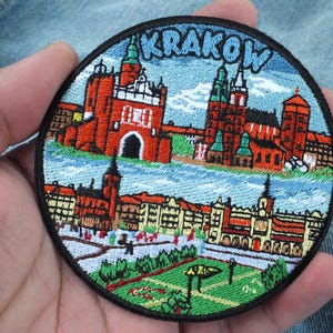 May include: Embroidered patch depicting Krakow, Poland. The design shows detailed illustrations of buildings, a river, and the city name in blue. The patch is round with a black border.