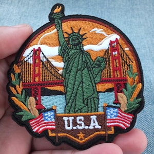 Może przedstawiać: Haftowana naszywka przedstawiająca Statuę Wolności, most Golden Gate i amerykańskie flagi. Naszywka ma czarną obwódkę i napis "U.S.A." w kolorze białym. Tło jest pomarańczowe i niebieskie.