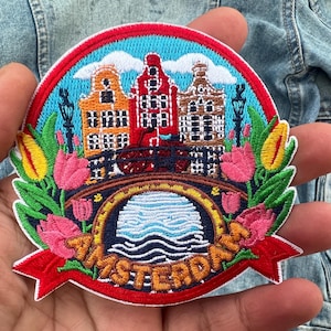 Op de afbeelding: Geborduurde patch met Amsterdamse grachtenhuizen, een brug en tulpen. Het ontwerp bevat het woord "Amsterdam" in een banner, tegen een blauwe lucht met wolken. De patch is rond met een rode rand.