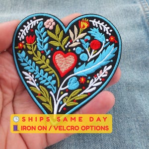 Może przedstawiać: Haftowana naszywka w kształcie serca z czarnym tłem. Naszywka przedstawia centralne czerwone serce, otoczone kolorowymi wzorami kwiatowymi i liściastymi w kolorze niebieskim, zielonym, białym i czerwonym. Naszywka ma niebieską obwódkę.