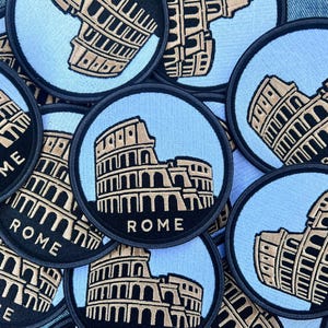 Può includere: Toppe rotonde raffiguranti il Colosseo di Roma. Le toppe hanno uno sfondo azzurro chiaro con un bordo nero. Il Colosseo è raffigurato in beige e la parola "ROME" è stampata in bianco.