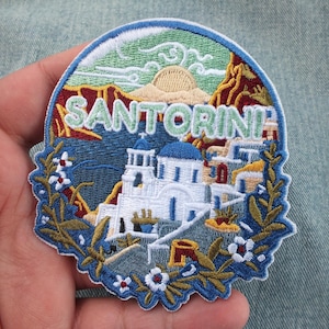 May include: Embroidered patch featuring the word "SANTORINI" above a depiction of the Greek island. The design includes white buildings with blue domes, a sunset, and floral accents. The patch is bordered with a blue and white scalloped edge.