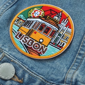 Puede incluir: Parche bordado con un tranvía amarillo de Lisboa, edificios y la palabra "LISBON" en una chaqueta vaquera. El parche tiene un diseño circular con un borde colorido y una bandera portuguesa.