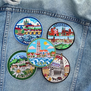 May include: Five embroidered patches featuring cityscapes. The patches are round with detailed designs of Prague, Krakow, Ulm, Salzburg, and another city. Each patch has a different color scheme, with buildings, water, and text identifying the city.