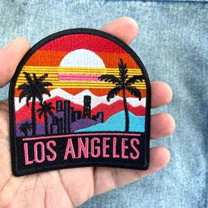 May include: Embroidered patch depicting a sunset over Los Angeles. The design includes palm trees, a cityscape silhouette, and the text "LOS ANGELES" in pink. The patch has a black border and a vibrant, colourful design.
