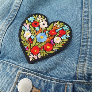 Könnte beinhalten: Ein herzförmiger, gestickter Aufnäher mit einem farbenfrohen Blumenmuster. Der Aufnäher zeigt rote, blaue, weiße und gelbe Blumen mit grünen Blättern auf schwarzem Grund. Der Aufnäher befindet sich auf einer Jeansjacke.