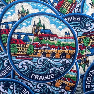 Puede incluir: Parche bordado con una detallada representación del paisaje urbano de Praga. El diseño incluye edificios icónicos, puentes y la palabra "PRAGUE" en letras blancas, rodeada por un borde azul y olas estilizadas.