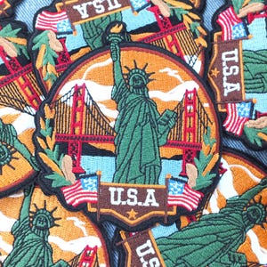 Peut inclure: Écussons brodés représentant la Statue de la Liberté et le Golden Gate Bridge. Les écussons sont circulaires avec une bordure marron et incluent le texte "U.S.A." et des drapeaux américains. Le fond est un mélange d'orange, de bleu et de vert.
