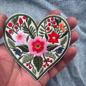 Puede incluir: Parche bordado en forma de corazón con un diseño floral. El parche tiene un fondo blanco y un borde verde. Las flores son rosas, rojas y naranjas, con hojas verdes y bayas rojas. El parche se sostiene en una mano.