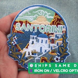 Peut inclure: Écusson brodé représentant l'île grecque de Santorin, avec le mot "SANTORINI" au-dessus d'une scène de bâtiments blancs aux dômes bleus, de montagnes et d'un coucher de soleil. Bordure bleue et détails floraux.