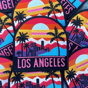 May include: Embroidered patch featuring a sunset scene with palm trees, mountains, and a city skyline. The patch has a black border and the text "LOS ANGELES" in pink. The background colors include red, orange, yellow, blue, and purple.