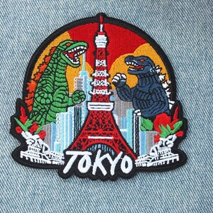 Peut inclure: Écusson brodé représentant des monstres Godzilla encadrant la Tour de Tokyo. L'écusson a une bordure noire, avec le mot "TOKYO" en blanc. Le fond comprend un paysage urbain et un ciel rouge, orange et jaune.