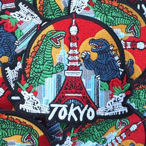 Peut inclure: Écussons brodés représentant Godzilla se battant devant la Tour de Tokyo. Les écussons sont rouges, jaunes et bleus, avec le mot "TOKYO" en blanc. Ces écussons sont un accessoire amusant.