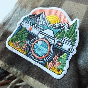 Peut inclure: Patch brodé représentant un appareil photo vintage avec un paysage de montagne et un coucher de soleil en arrière-plan. L'appareil photo est blanc avec un objectif noir et un bouton rouge. Les montagnes sont vertes et bleues avec un coucher de soleil rose et jaune. Le patch est entouré d'une bordure blanche.
