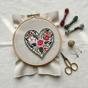 Op de afbeelding: Een borduurring met een hartvormig bloemenontwerp. Het hart heeft roze, rode en witte bloemen met groene bladeren en rode bessen. Borduurgaren, een vingerhoed, naalden en een kleine gouden schaar zijn ook aanwezig.