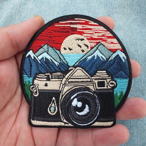 May include: Embroidered patch with a vintage camera design set against a backdrop of mountains, a lake, and a sunset. The patch is circular, with a black border and detailed stitching in red, blue, and beige.