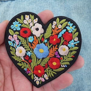 May include: Embroidered heart-shaped patch featuring a colourful floral design. The patch features red, blue, white and yellow flowers with green leaves on a black background.