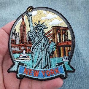 Può includere: Toppa ricamata con una rappresentazione colorata della Statua della Libertà, il ponte di Brooklyn e lo skyline di New York. La toppa ha uno stendardo blu con il testo "NEW YORK" in bianco.