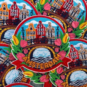 May include: Embroidered patch featuring Amsterdam canal houses, a bridge, and tulips. The design includes the word "Amsterdam" on a red banner, set against a blue sky and water background. The patch is surrounded by green leaves and colorful flowers.