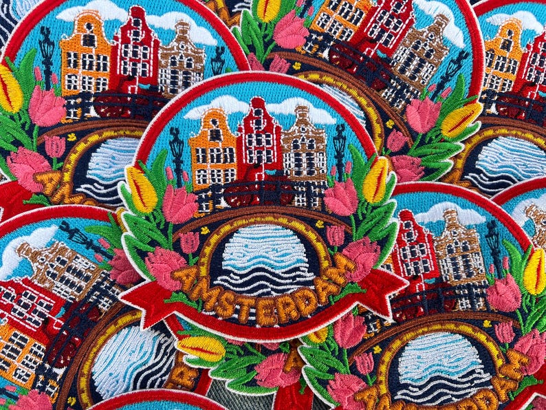 May include: Embroidered patch featuring Amsterdam canal houses, a bridge, and tulips. The design includes the word "Amsterdam" on a red banner, set against a blue sky and water background. The patch is surrounded by green leaves and colorful flowers.