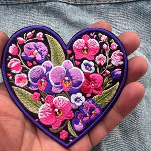 Puede incluir: Parche bordado en forma de corazón con un diseño floral. Presenta orquídeas rosas, moradas y blancas y otras flores, con hojas verdes sobre un fondo negro. El parche está delineado en morado oscuro.