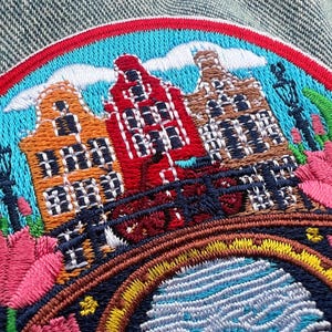 May include: Embroidered patch featuring a colorful depiction of Dutch canal houses, a bridge, and tulips. The patch has a red, blue, and yellow border. The houses are orange, red, and brown. The patch is on a denim background.