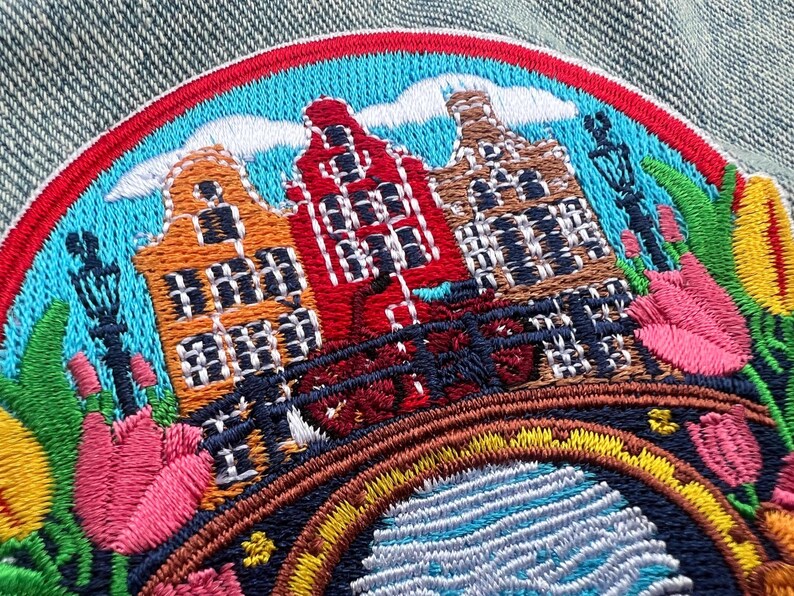 May include: Embroidered patch featuring a colorful depiction of Dutch canal houses, a bridge, and tulips. The patch has a red, blue, and yellow border. The houses are orange, red, and brown. The patch is on a denim background.