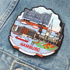 May include: Embroidered patch depicting Hamburg, Germany, with the city's name in red. The patch shows buildings, boats, and water, set against a light blue background, sewn onto a denim jacket.