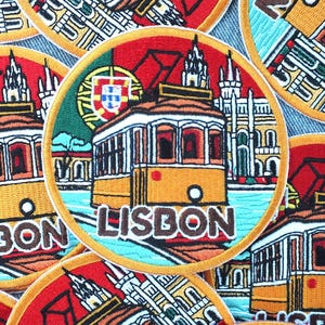 Puede incluir: Parches bordados circulares que representan un tranvía amarillo de Lisboa, con el nombre de la ciudad en blanco. Los parches tienen un borde rojo y naranja, con un fondo verde y azul. El diseño incluye elementos arquitectónicos y la bandera portuguesa.