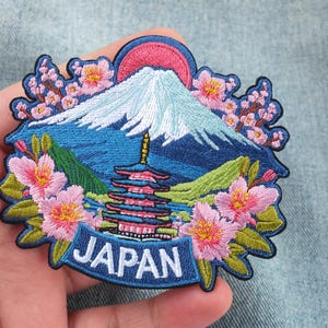 Peut inclure: Écusson brodé représentant le Mont Fuji, une pagode et des fleurs de cerisier. Le motif comprend un ciel bleu, des collines vertes et un soleil rose. Le mot "JAPAN" est affiché sur une bannière bleue.