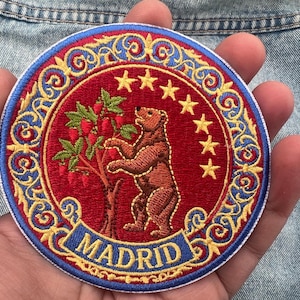 Puede incluir: Parche bordado redondo con un oso pardo que alcanza bayas rojas en un árbol, rodeado de estrellas doradas y un borde decorativo. La palabra "MADRID" se muestra en una pancarta azul.