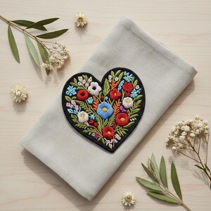 May include: A heart-shaped embroidered patch with a black border, featuring a vibrant floral design in red, blue, white, and yellow. It's placed on a folded grey linen napkin, with sprigs of greenery and dried flowers around it.