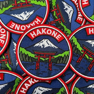 May include: A circular embroidered patch featuring a landscape with a snow-capped mountain, a red torii gate in blue water, and green trees. The word HAKONE is stitched in white letters on a red curved banner at the top of the circular design.