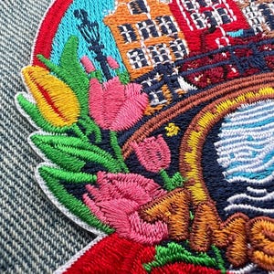 May include: Embroidered patch featuring a colorful design of tulips, buildings, and a bridge. The patch has a circular shape with a red, blue, and yellow border. The buildings are orange, red, and brown. The tulips are pink, yellow, and red.