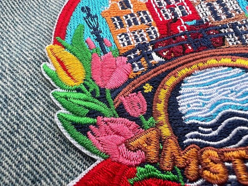 May include: Embroidered patch featuring a colorful design of tulips, buildings, and a bridge. The patch has a circular shape with a red, blue, and yellow border. The buildings are orange, red, and brown. The tulips are pink, yellow, and red.