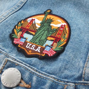 Könnte beinhalten: Gestickter Aufnäher mit der Freiheitsstatue, der Golden Gate Bridge und dem Schriftzug "U.S.A." in einem runden Design mit roten, weißen und blauen Akzenten.