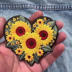 Könnte beinhalten: Gestickter herzförmiger Aufnäher mit drei großen Sonnenblumen mit gelben Blütenblättern und roten Zentren, umgeben von kleineren gelben Blüten und grünen Blättern. Der Aufnäher befindet sich auf einem dunkelbraunen Hintergrund.