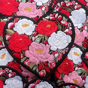 May include: Embroidered heart-shaped patches featuring a floral design. The patches showcase red, pink, and white flowers with yellow centers, set against a black background. The patches are ideal for adding a decorative touch to clothing or accessories.