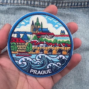 May include: A circular embroidered patch showing a colourful illustration of Prague, Czech Republic. The design includes buildings, a bridge, water, and the word "PRAGUE" in white lettering on a blue background.