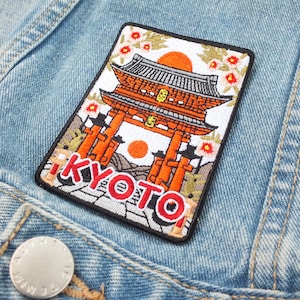 May include: Embroidered patch featuring a Japanese temple with orange and white architectural details, a rising sun, and the word "KYOTO" in red. The patch is rectangular with a black border and is sewn onto a light blue denim jacket.
