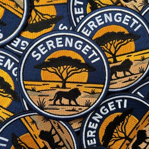 May include: A collection of circular embroidered patches featuring a Serengeti landscape. Each patch displays a dark blue border with the text SERENGETI in white, a yellow sun, a black acacia tree silhouette, and a black lion standing on grass.