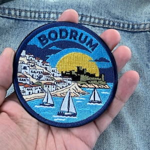 May include: A circular embroidered patch featuring a coastal scene with the text BODRUM at the top. The design includes white buildings with red roofs, a yellow sun, a dark fortress, and three sailboats on blue water, all bordered by a dark blue edge.