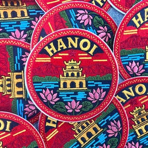 Puede incluir: Parches bordados con un diseño circular, con la palabra "HANOI" en letras doradas. Los parches representan una pagoda, flores de loto y un puente sobre un fondo rojo. Los parches son una vibrante mezcla de colores.
