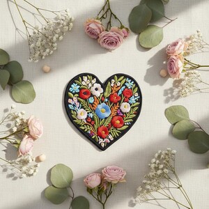 Op de afbeelding: Een hartvormige geborduurde patch met een zwarte rand, gevuld met kleurrijke bloemen in rood, blauw, wit en geel. De patch is omgeven door roze rozen, gipskruid en groene bladeren, wat een bloemstuk creëert.