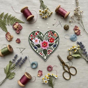 May include: A heart-shaped embroidered patch with a floral design, surrounded by sewing supplies. The patch features pink, red, and white flowers with green leaves. Spools of thread, scissors, and dried flowers are scattered around the patch.